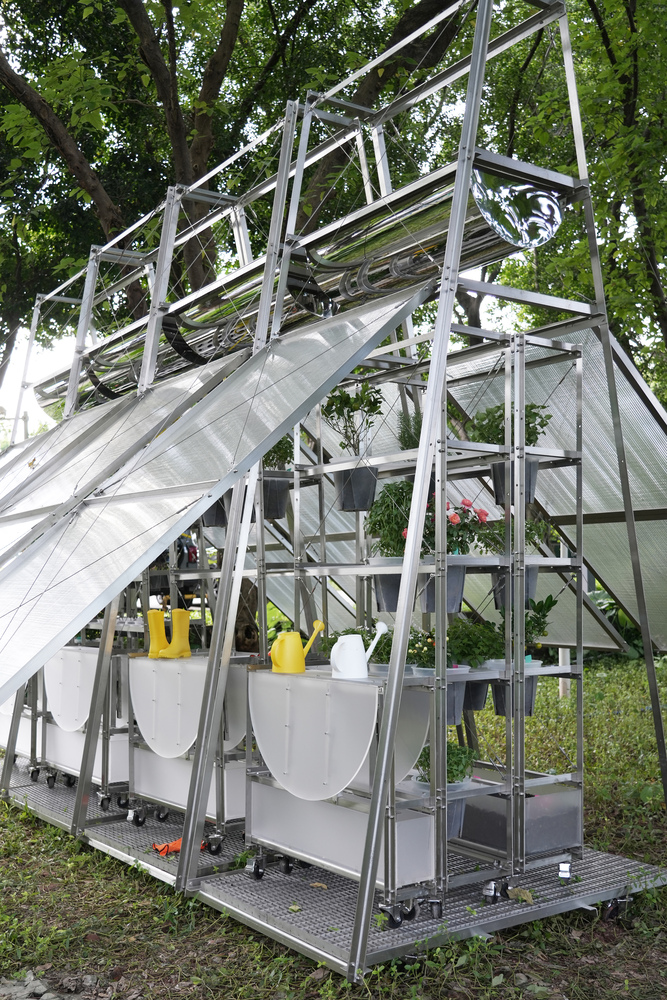 Angled steel frame greenhouse with translucent polycarbonate panels and hanging planters beneath a leafy canopy