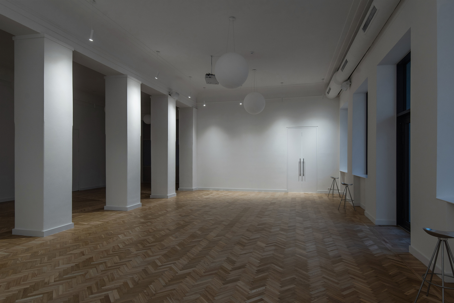 Open room with white columns framing doorways, herringbone wood flooring and sphere pendant lights overhead