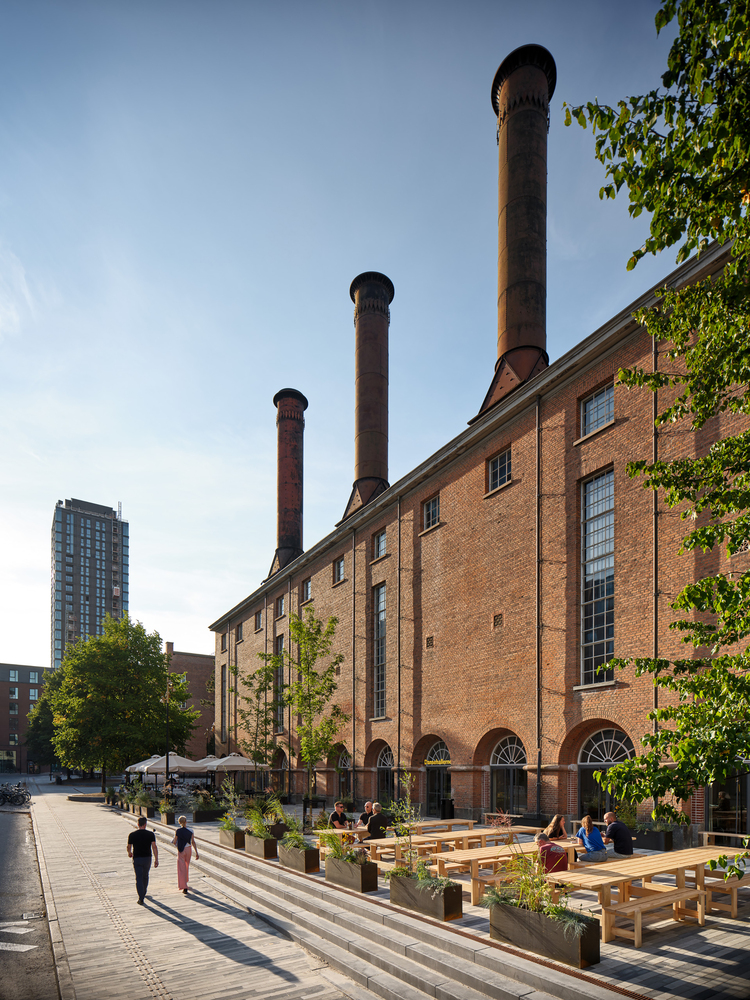 Outdoor seating area with timber benches and planters facing the brick facade with three chimneys