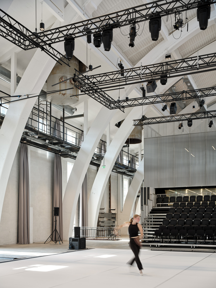Performance hall interior with white branching columns supporting stage lighting trusses as a figure walks across the floor