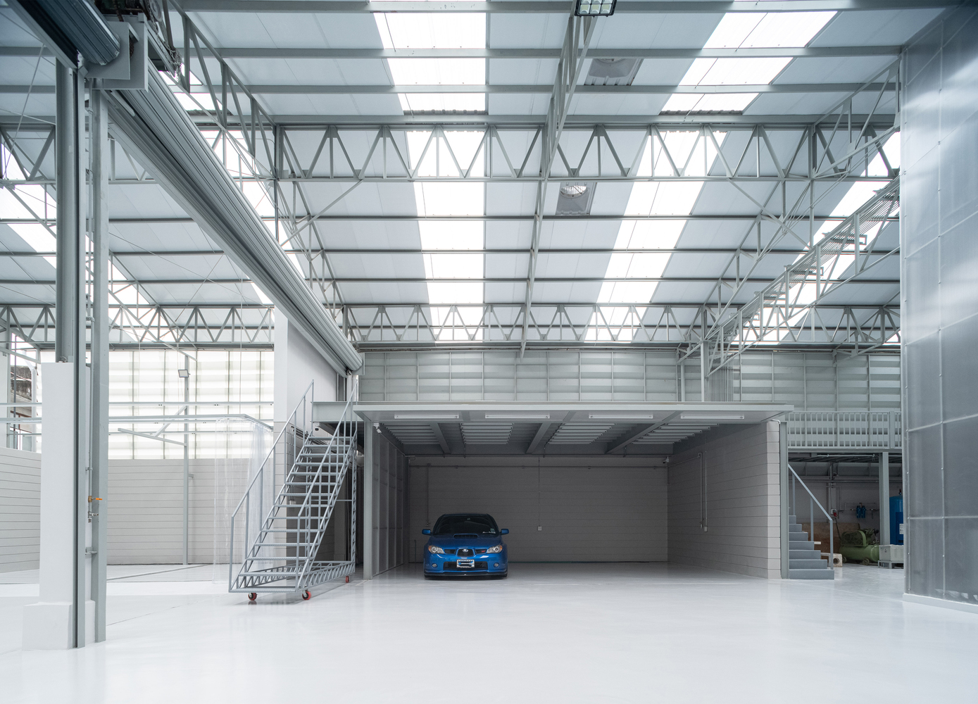 Interior warehouse space with exposed steel truss ceiling, mezzanine level, and blue car on white floor
