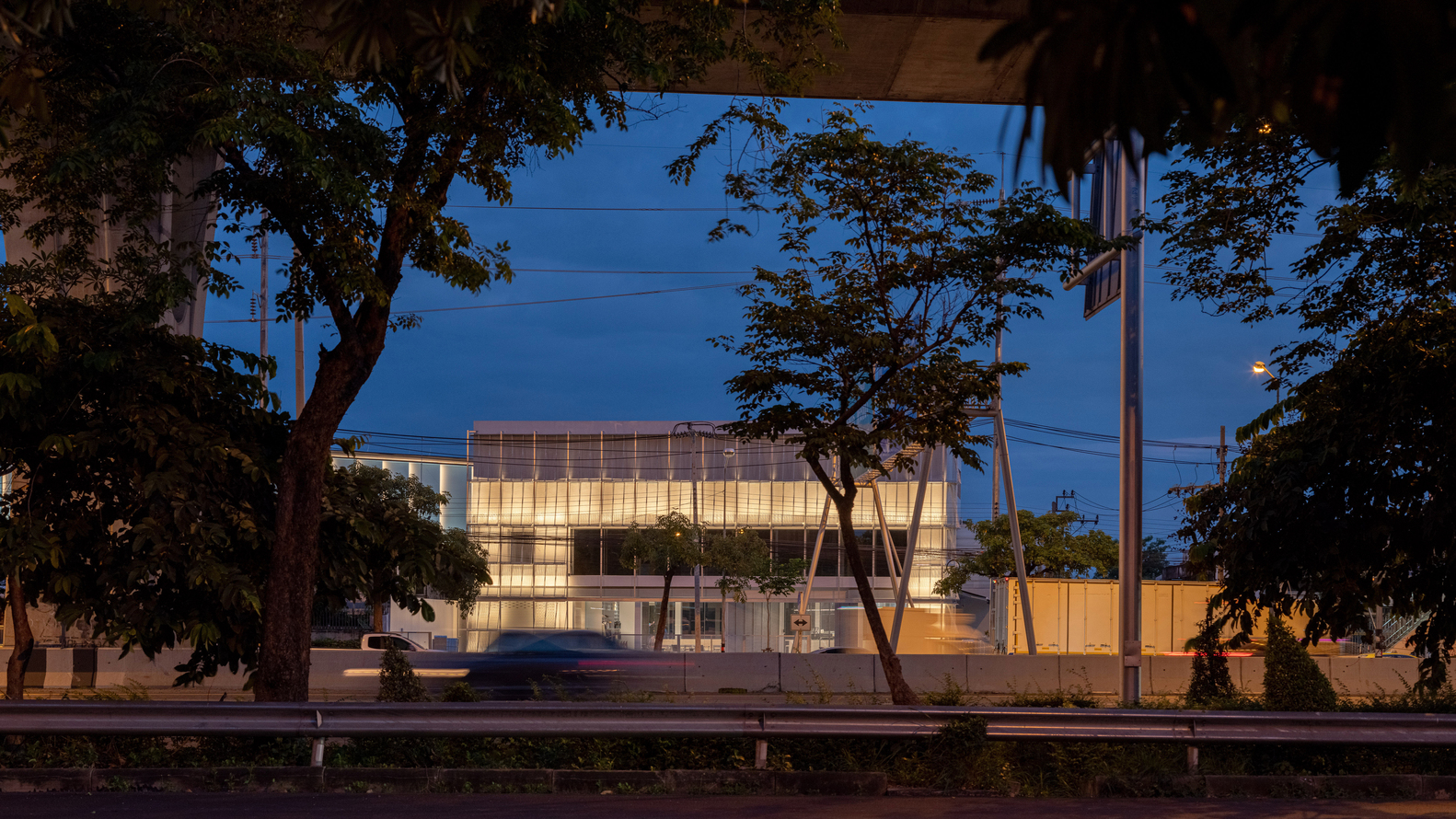Street view of the illuminated translucent facade framed by mature trees at dusk