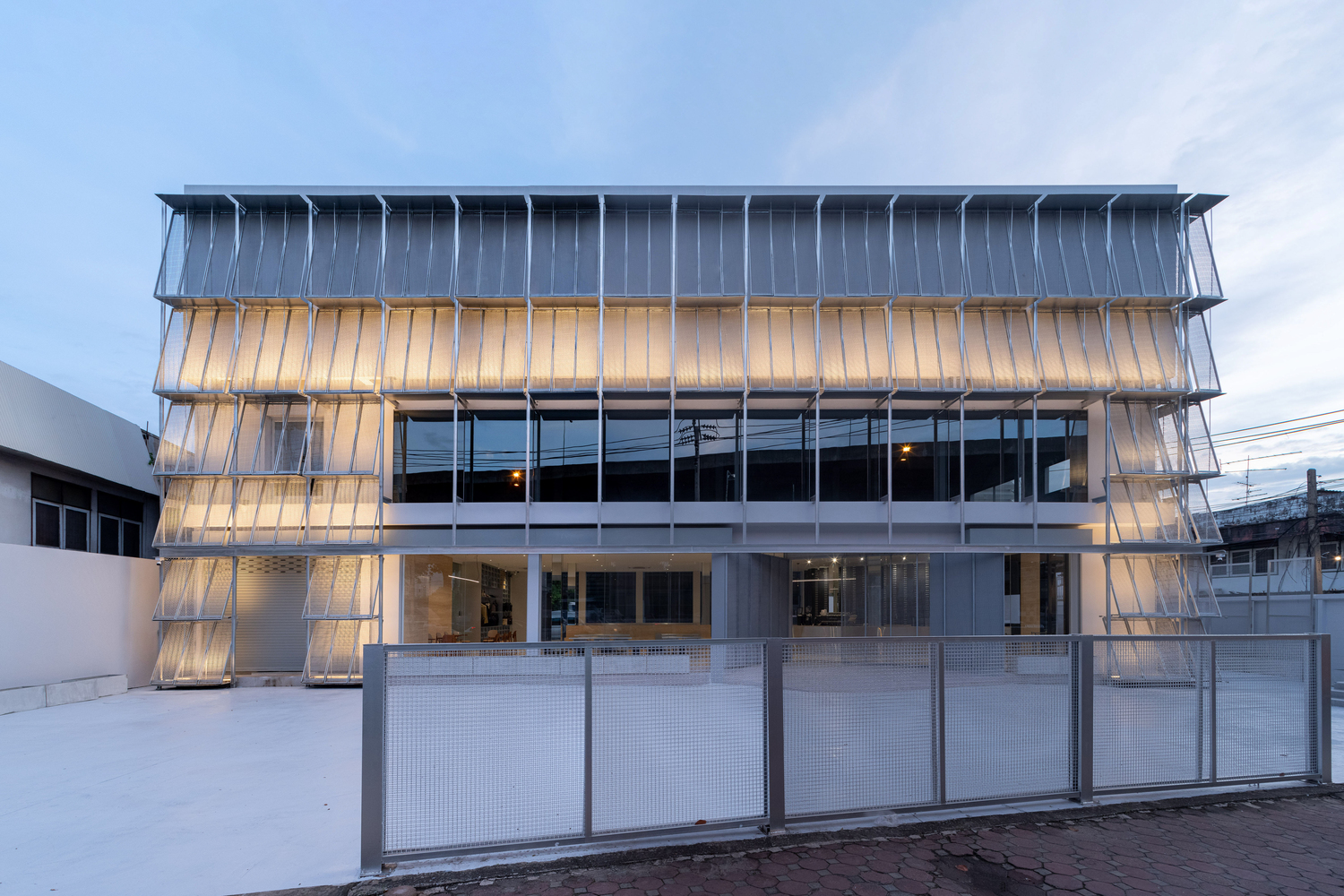 Layered translucent screen facade with white perforated metal fence in front at twilight