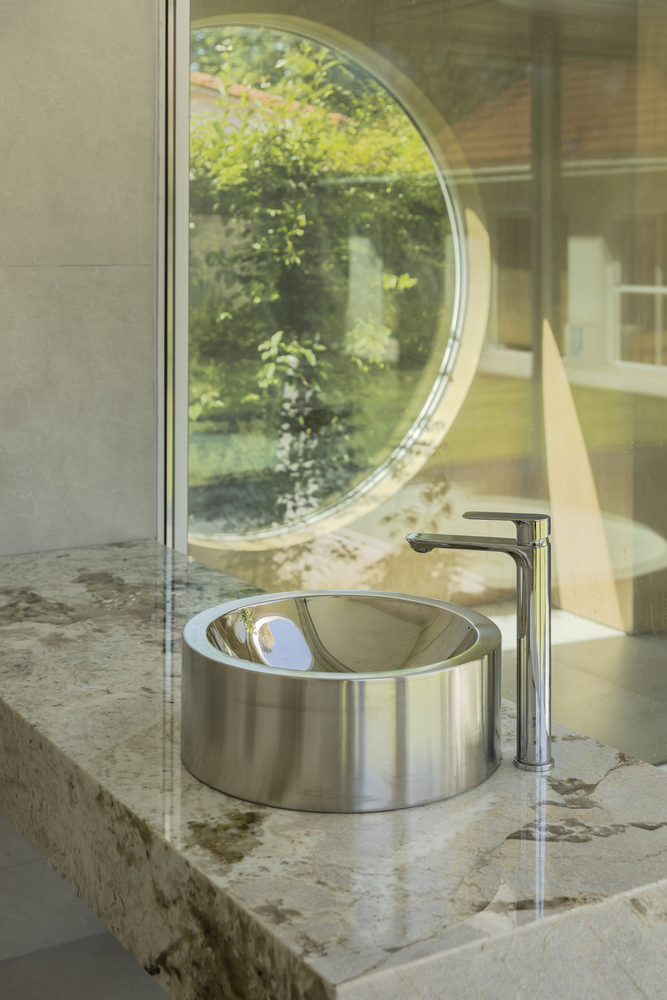 Stainless steel basin on stone countertop reflecting the circular garden window behind glass