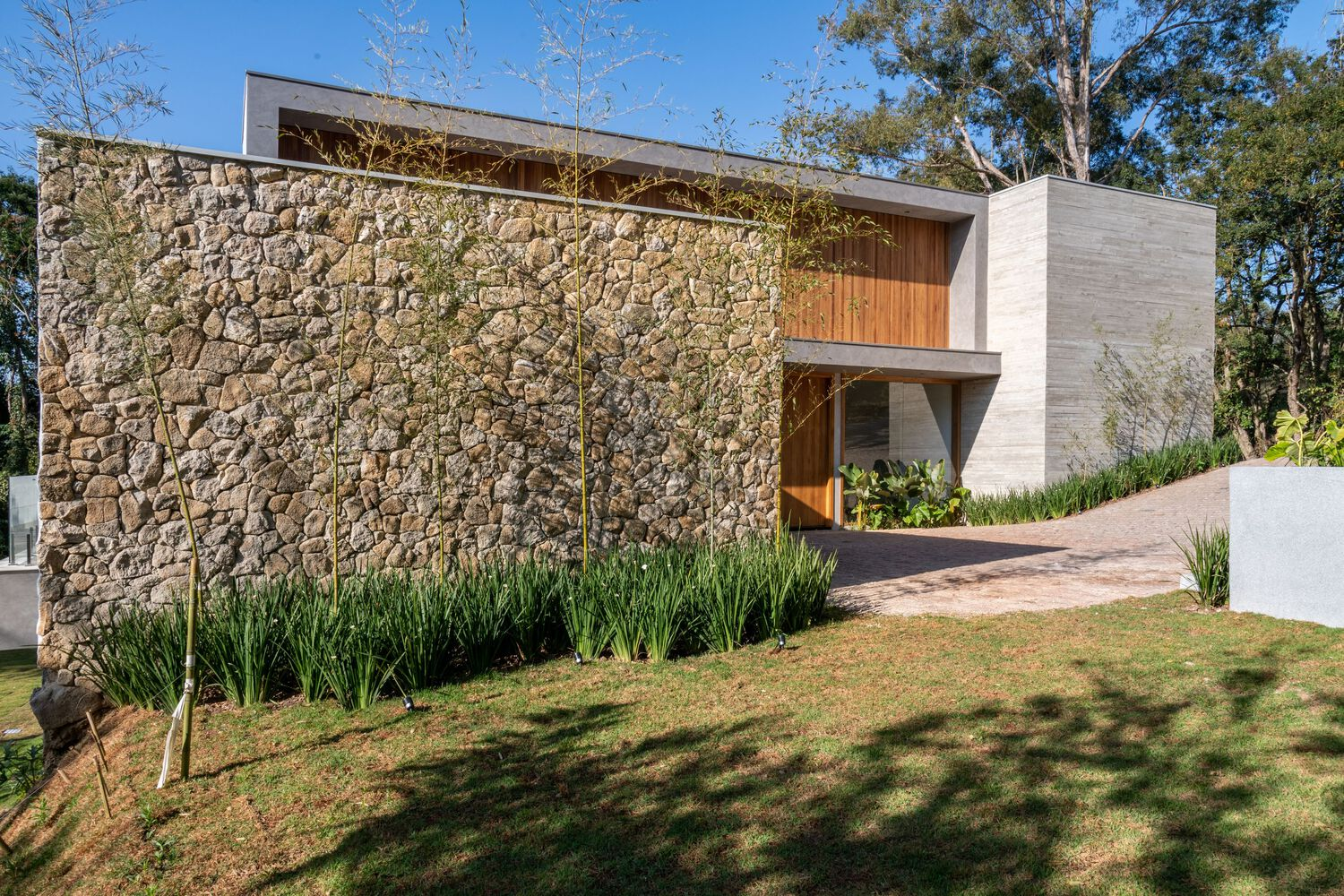 Entry facade with rough stone retaining wall, timber cladding and planted border along sloped drive