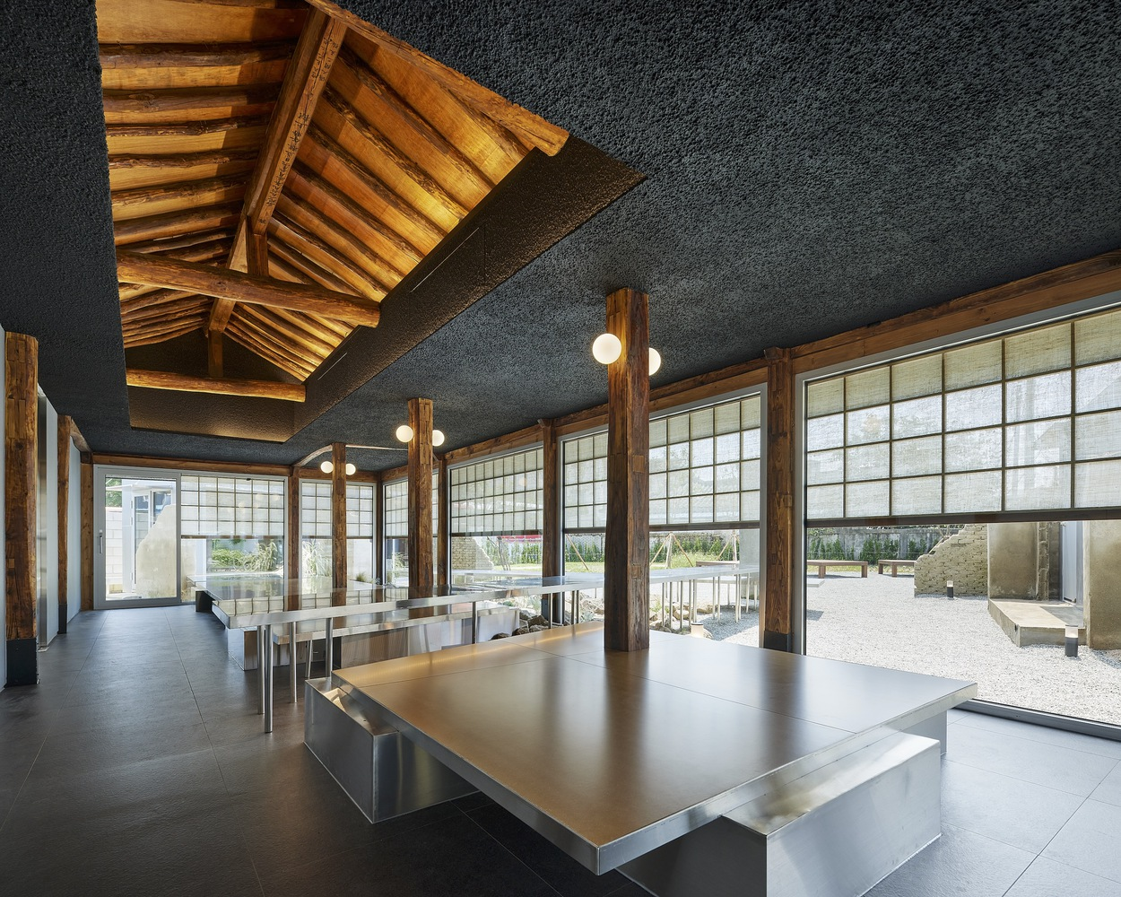 Interior hall with timber roof structure, translucent grid windows, and stainless steel tables on dark flooring