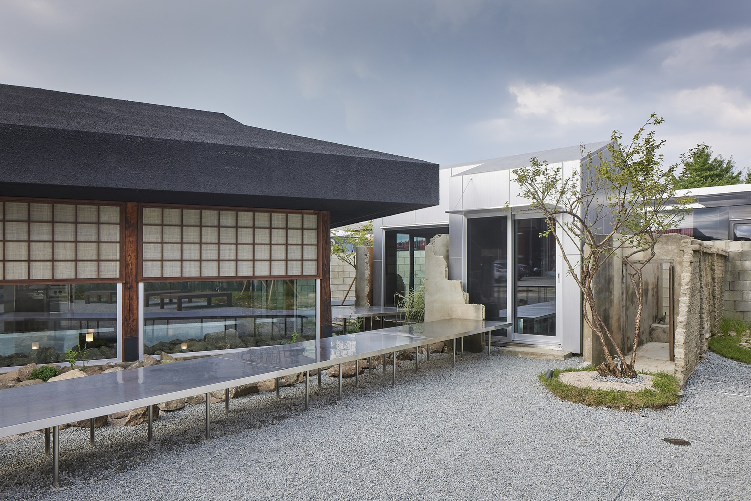 Gravel courtyard showing the pavilion with gridded facade, elevated steel walkway, and planted tree at dusk