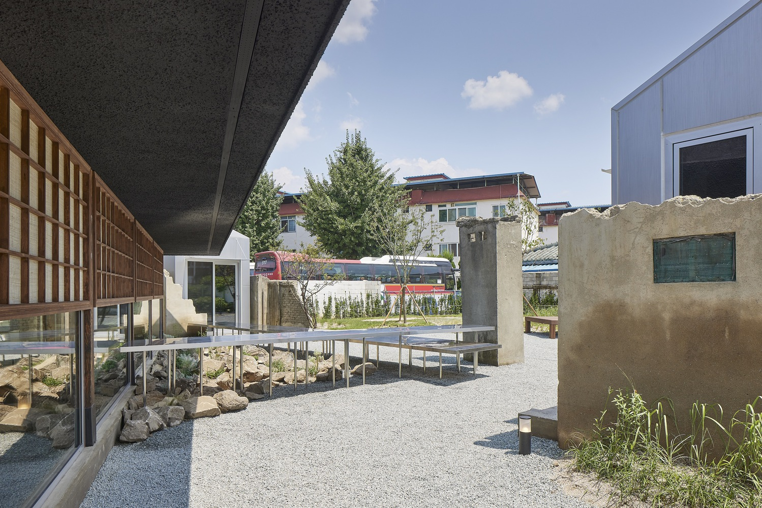 Gravel courtyard with metal benches, planted beds, and gridded timber screens under a blue sky