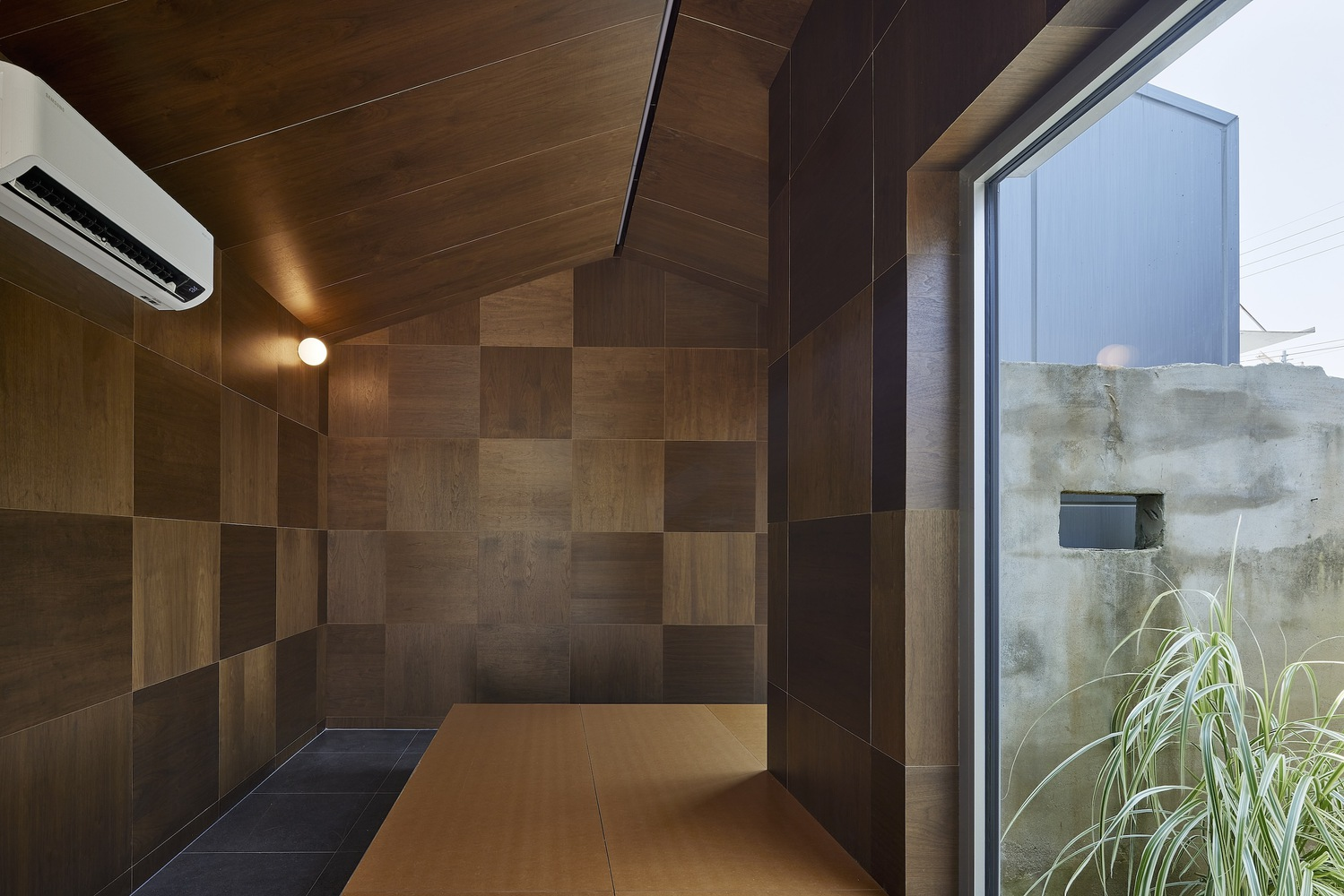 Room with chequered wood paneling on walls and ceiling and view to a concrete wall
