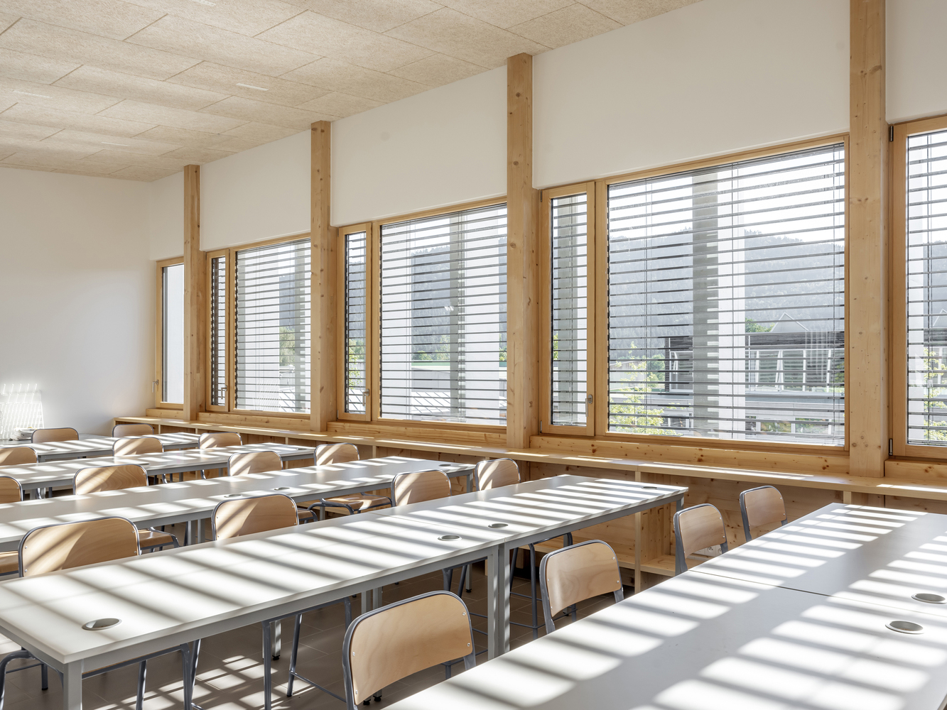Classroom interior with timber columns and row of windows casting slatted shadows across tables
