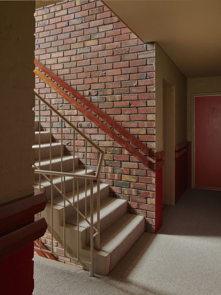 Internal staircase with steel and timber railing against an exposed brick wall lit by slanted sunlight