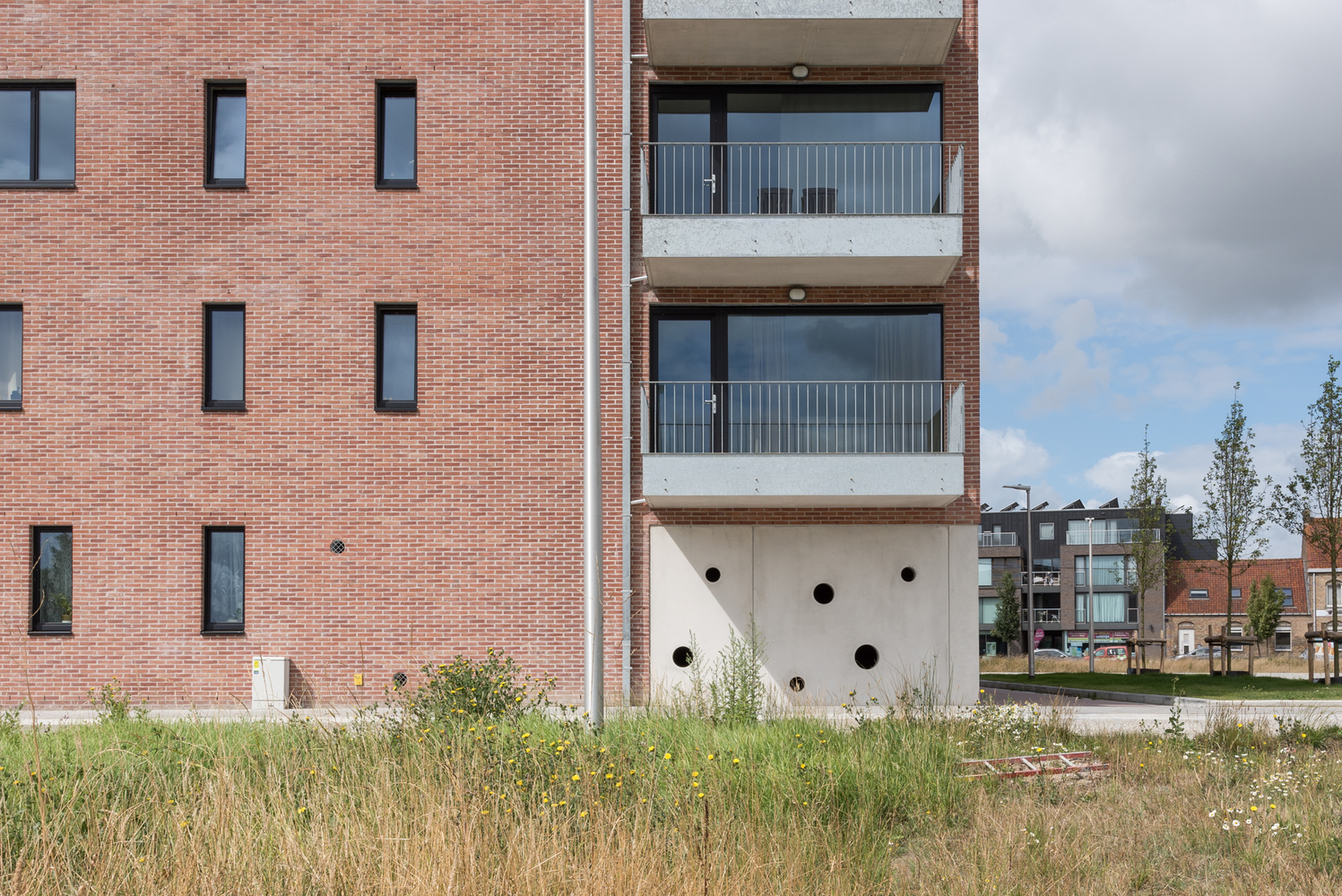 White concrete base with circular openings beneath stacked balconies and red brick facade in summer grasses