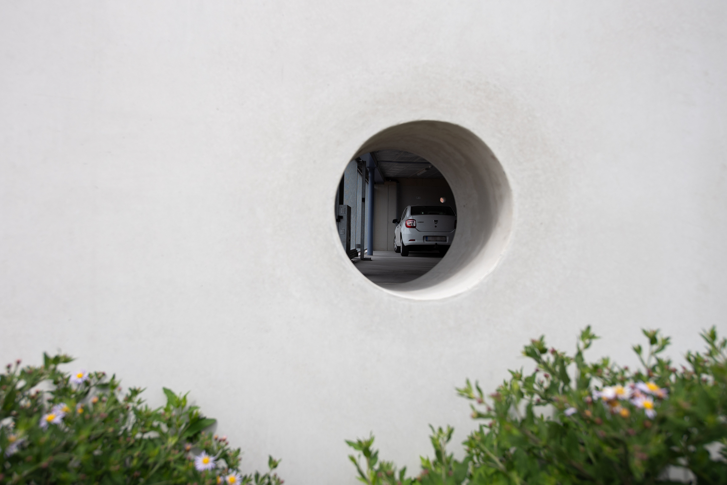 Circular opening in white wall revealing parked car beyond, with flowering shrubs below