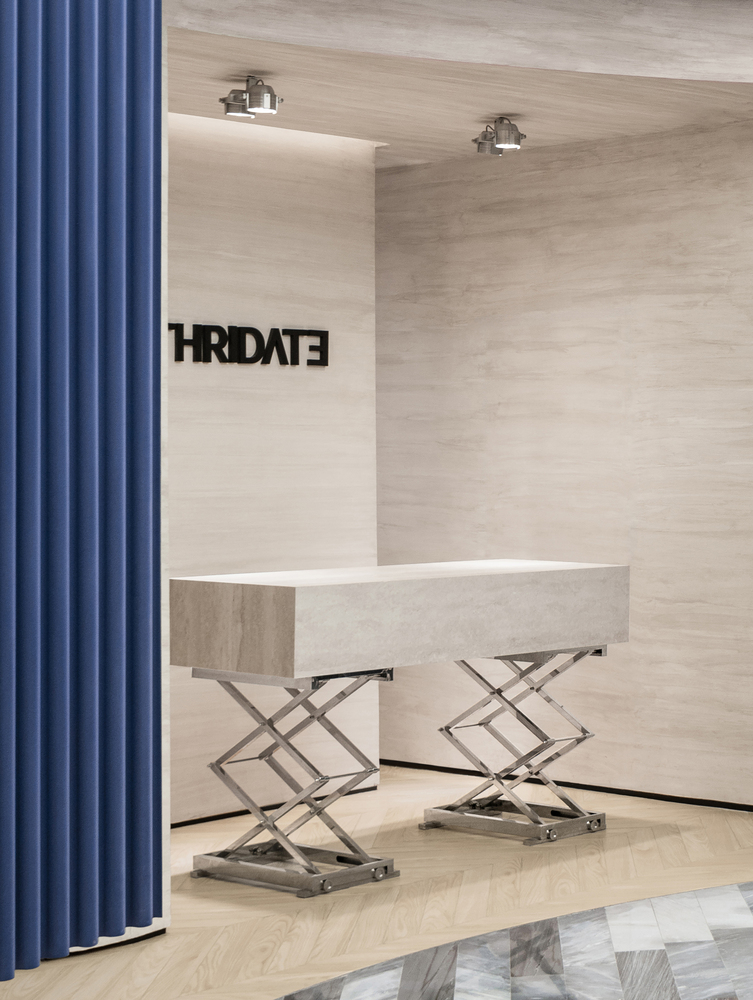 Reception desk on scissor-lift bases against travertine wall with blue curtain at left