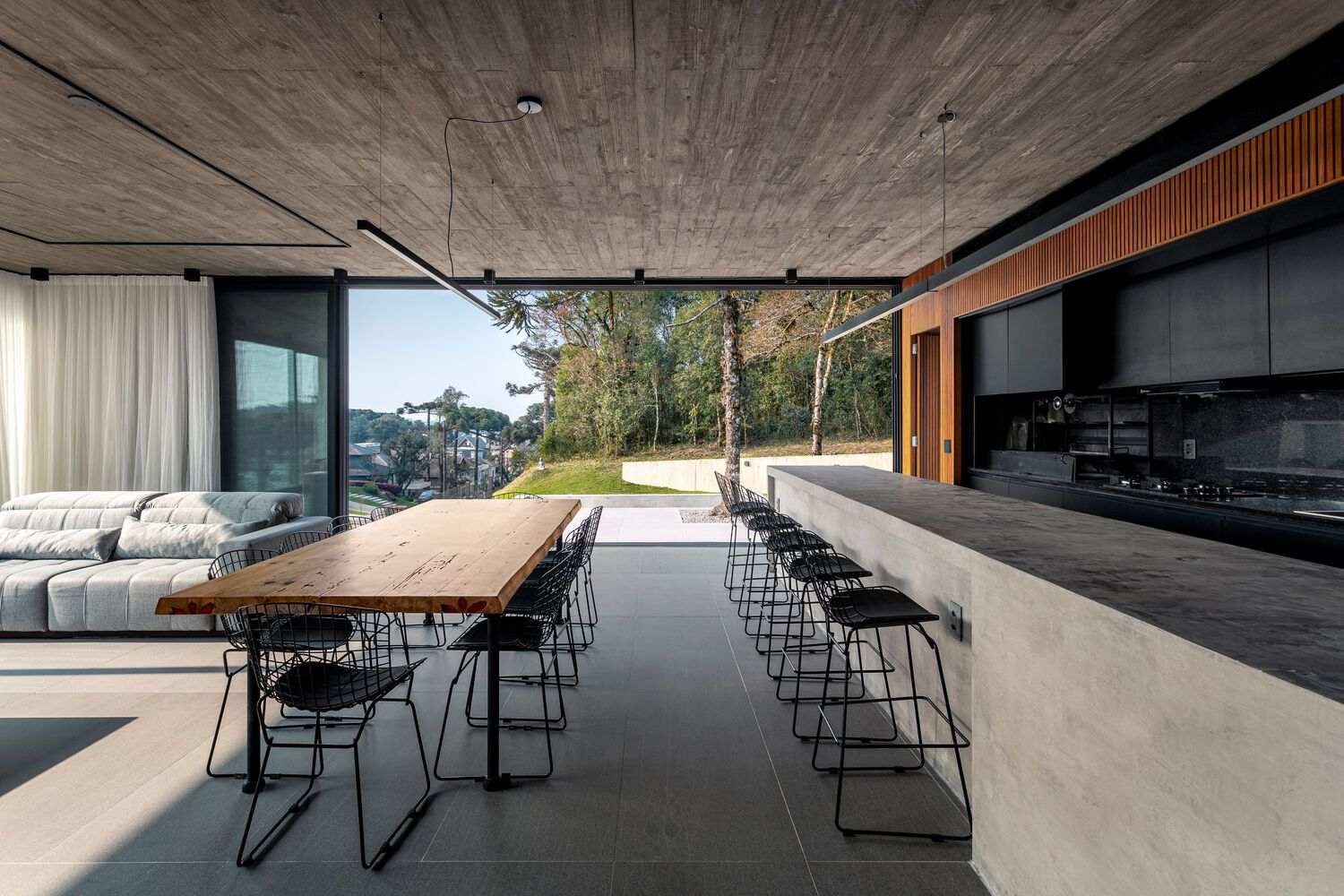 Open living space with board-formed concrete ceiling, live-edge dining table and black metal chairs opening to lawn