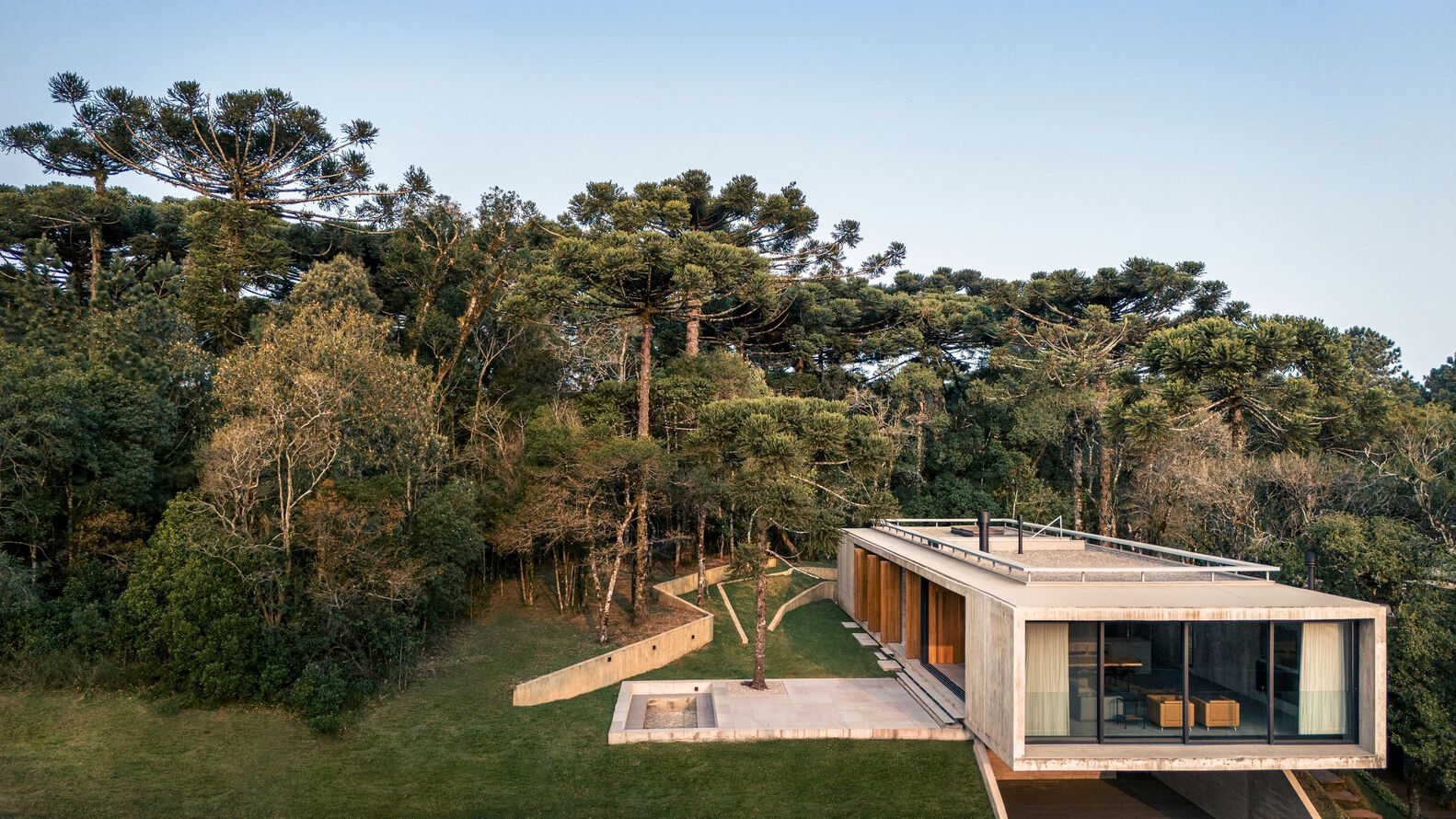 Elevated concrete structure with timber screens and rooftop terrace nestled into a forested hillside at dusk