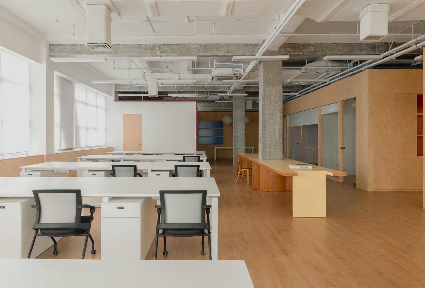 Open office space with exposed concrete columns, white desks and plywood partitions under a coffered ceiling