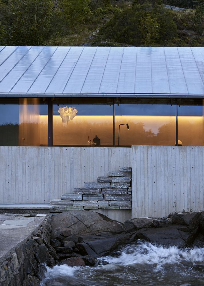 Glazed facade with board-formed concrete base and stone steps at water's edge at dusk