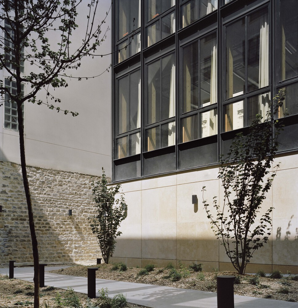 Gridded glass facade rising above stone base wall with young trees and climbing vines in the forecourt