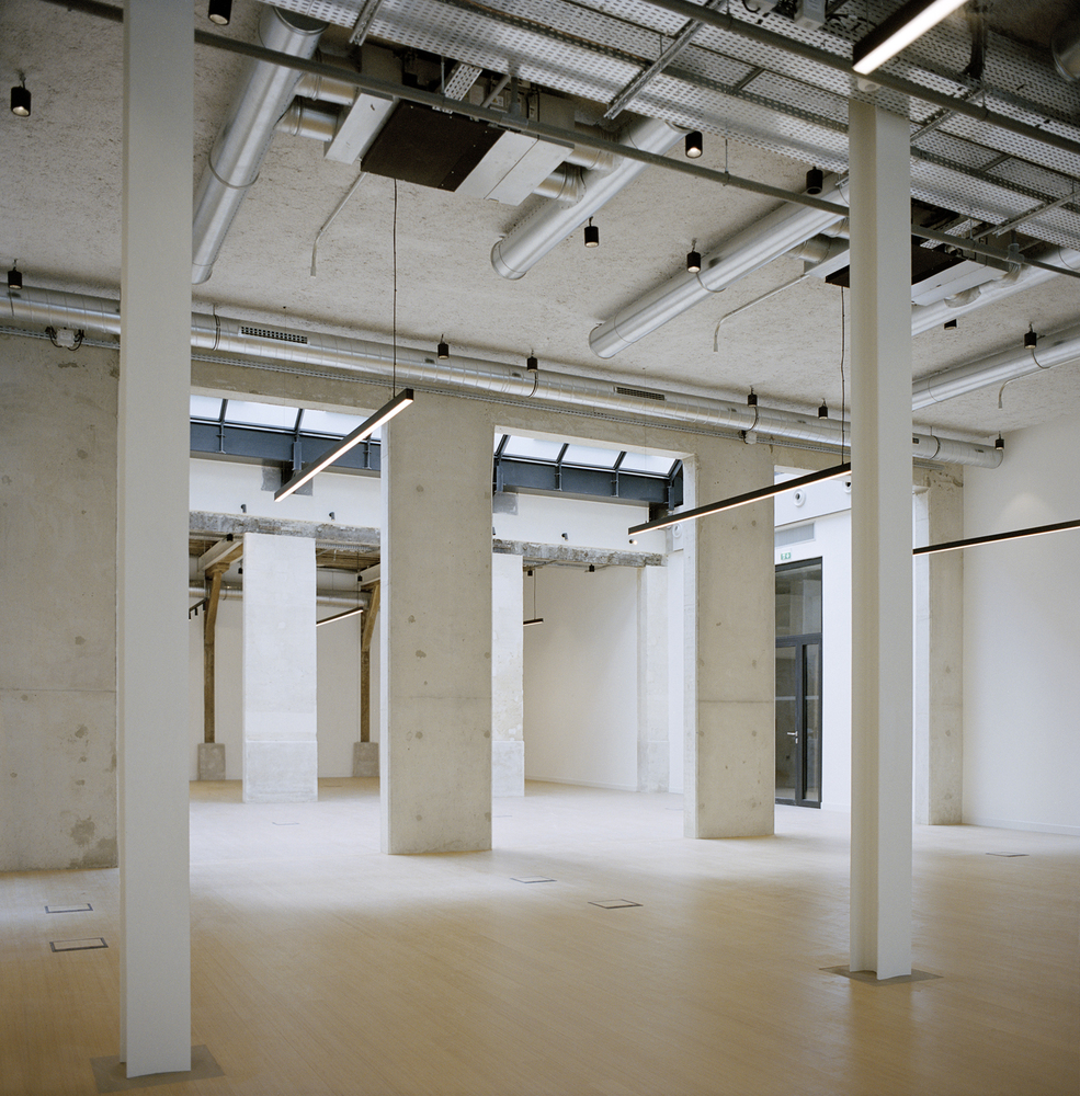Open interior space with exposed concrete columns and ceiling ducts beneath a perforated metal screen skylight