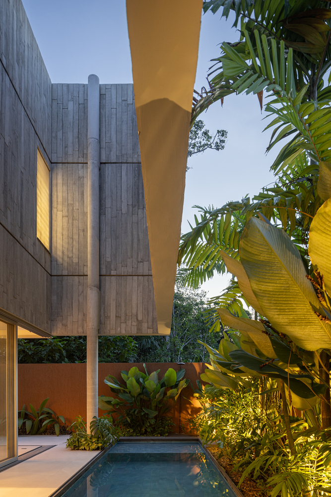 Courtyard pool framed by cantilevered yellow soffit and concrete pillar amid tropical plantings at dusk