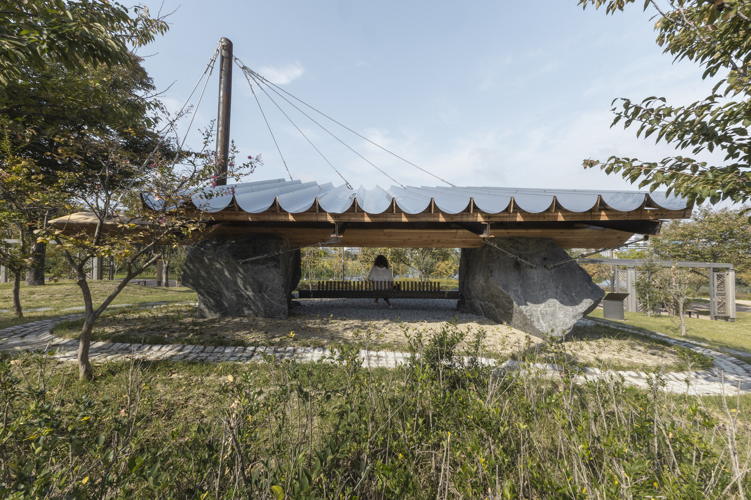 Open pavilion with undulating roof supported by large boulders and timber beams in a park setting