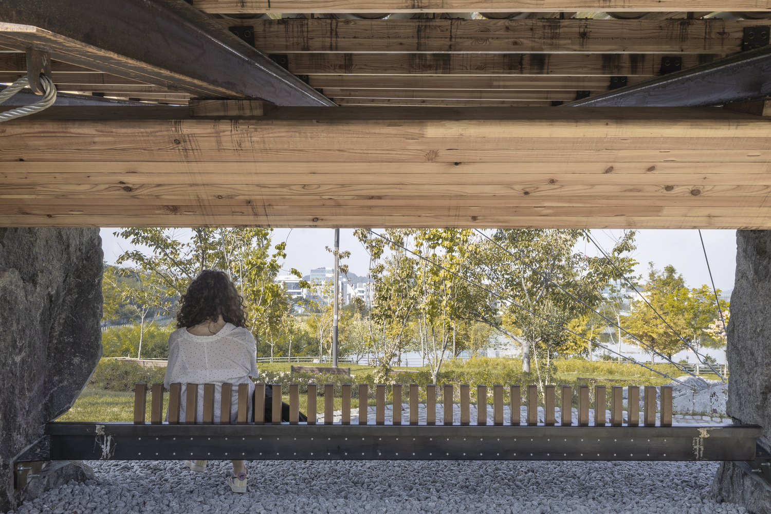 View through the pavilion showing a seated visitor and park landscape beyond the slatted wooden bench