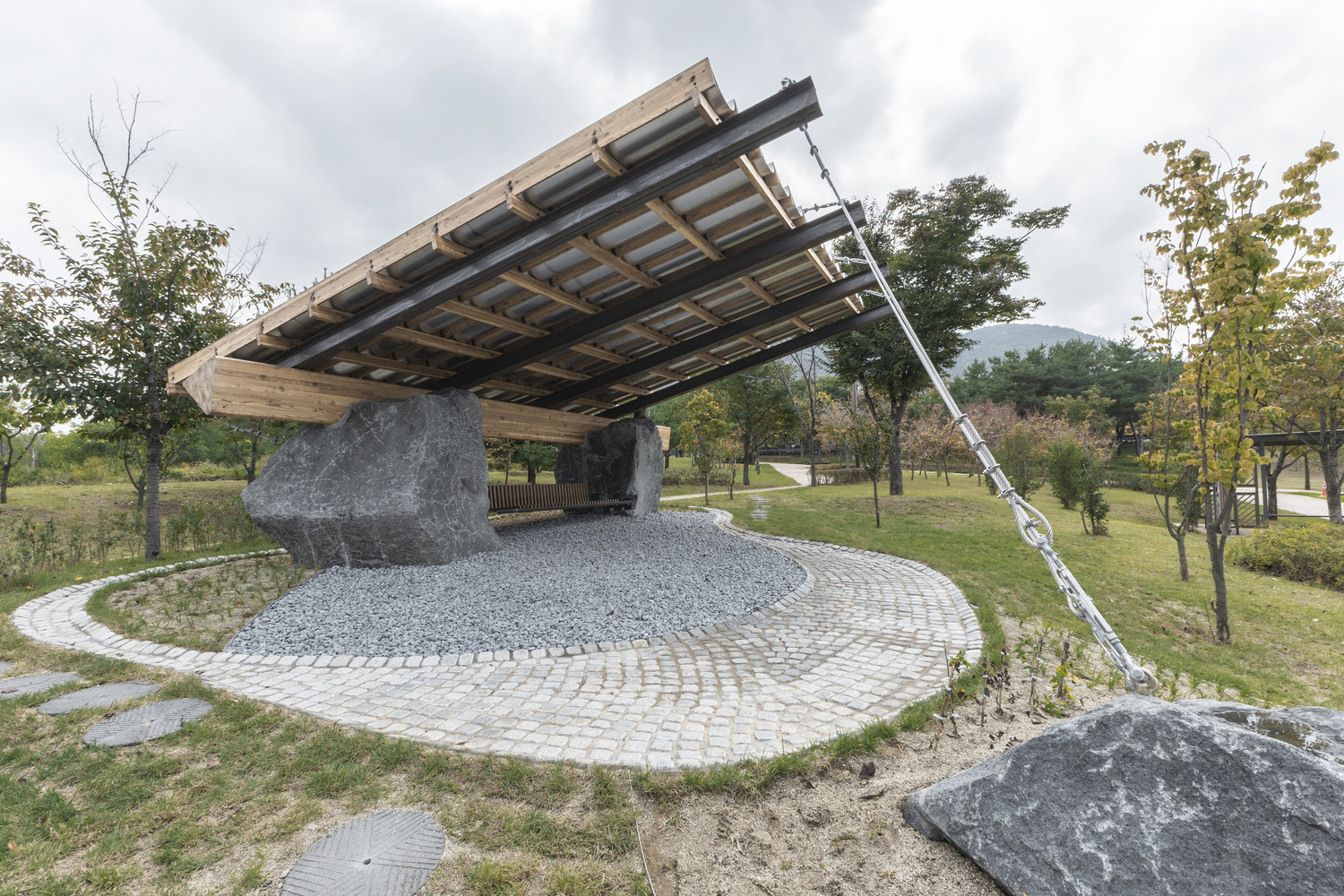 Tilted timber and steel canopy structure supported by large granite boulders over a circular paved platform