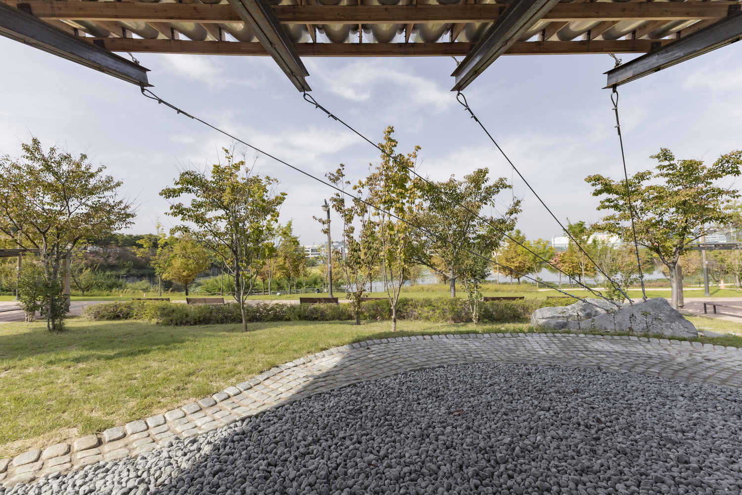 View from beneath the timber beam structure looking out toward a landscaped park with young trees