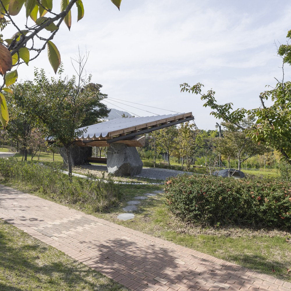 Angled pavilion roof supported by large boulders in a green park with brick pathways