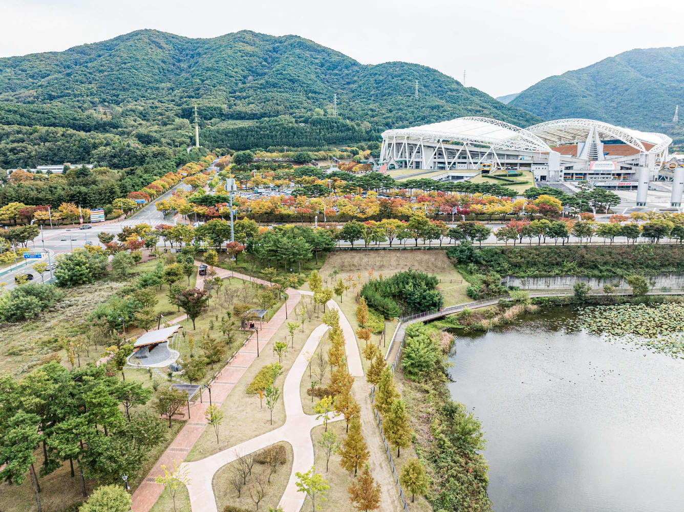 Elevated view of the riverside park with curving paths and a stadium complex beyond forested hills