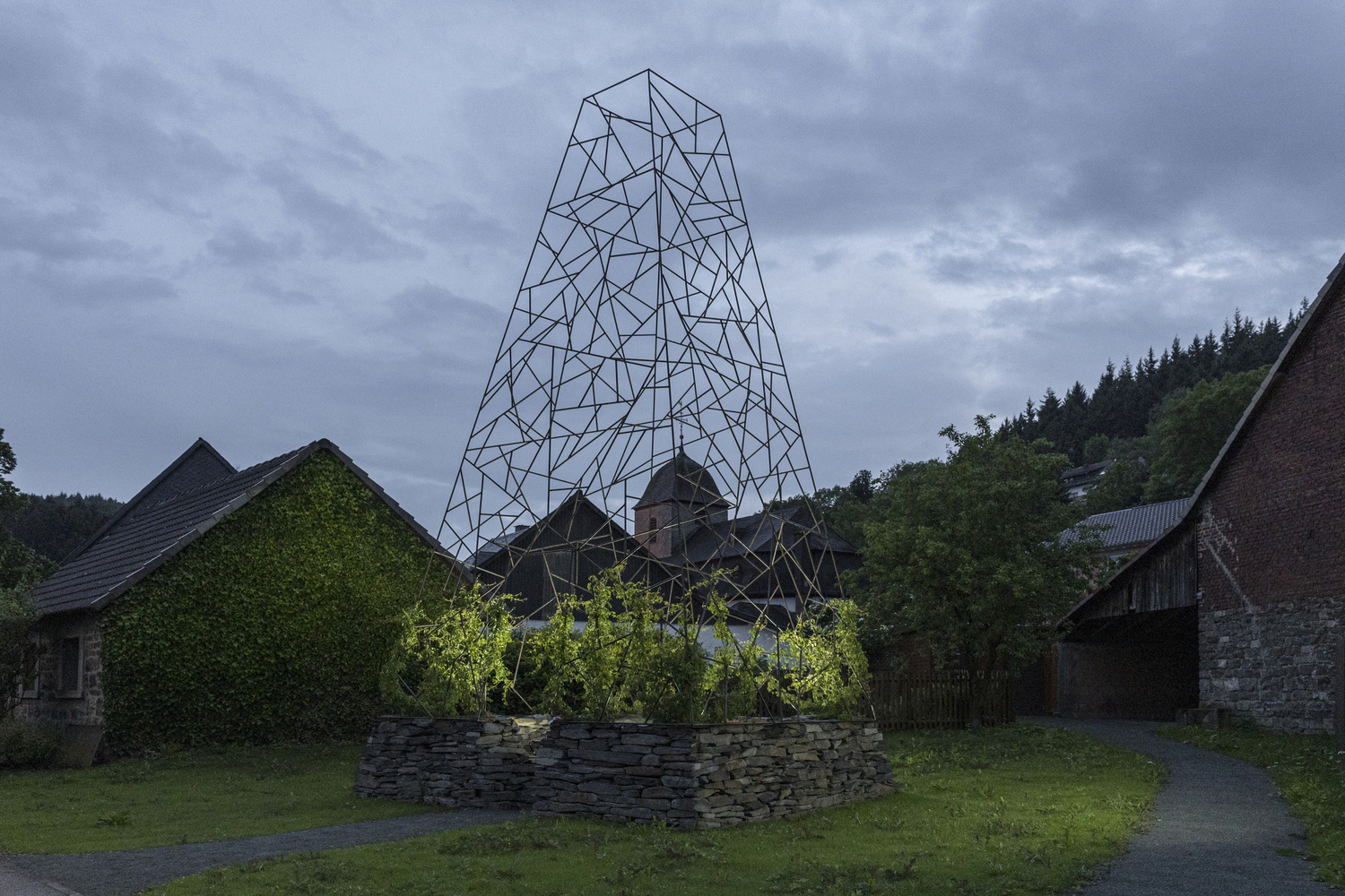 The wireframe tower and stone planting bed under an overcast evening sky beside rural farm buildings