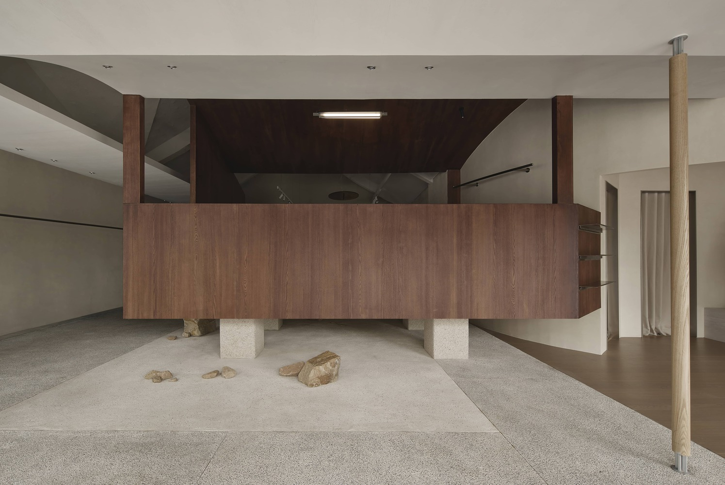 Walnut-clad volume elevated on square concrete blocks with loose stones scattered beneath on terrazzo