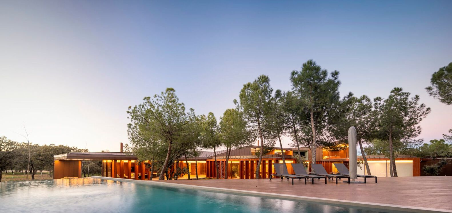 Pool terrace with timber decking and glazed volumes illuminated at twilight among scattered pine trees