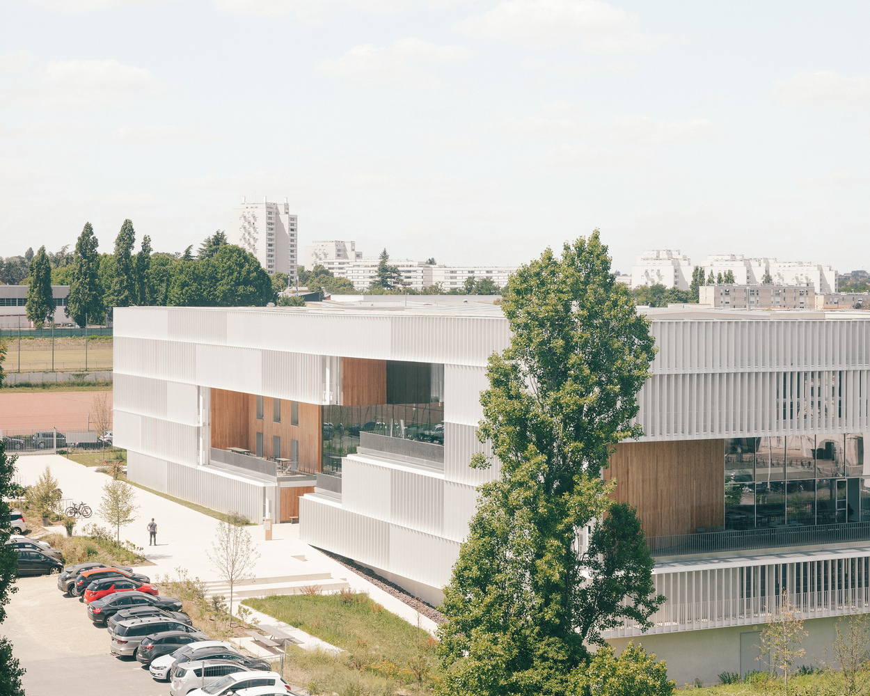 Elevated view of the white louvered facade with timber-clad volumes flanked by trees and parked cars