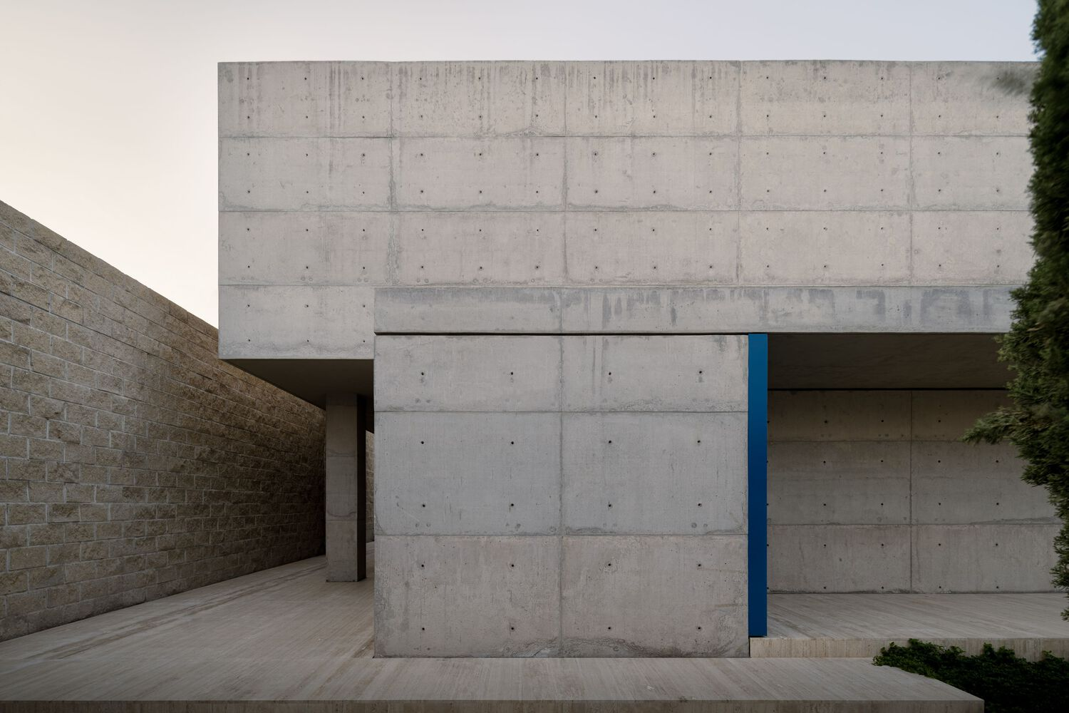 Board-formed concrete facade with recessed entry passage and single blue-painted panel at dusk