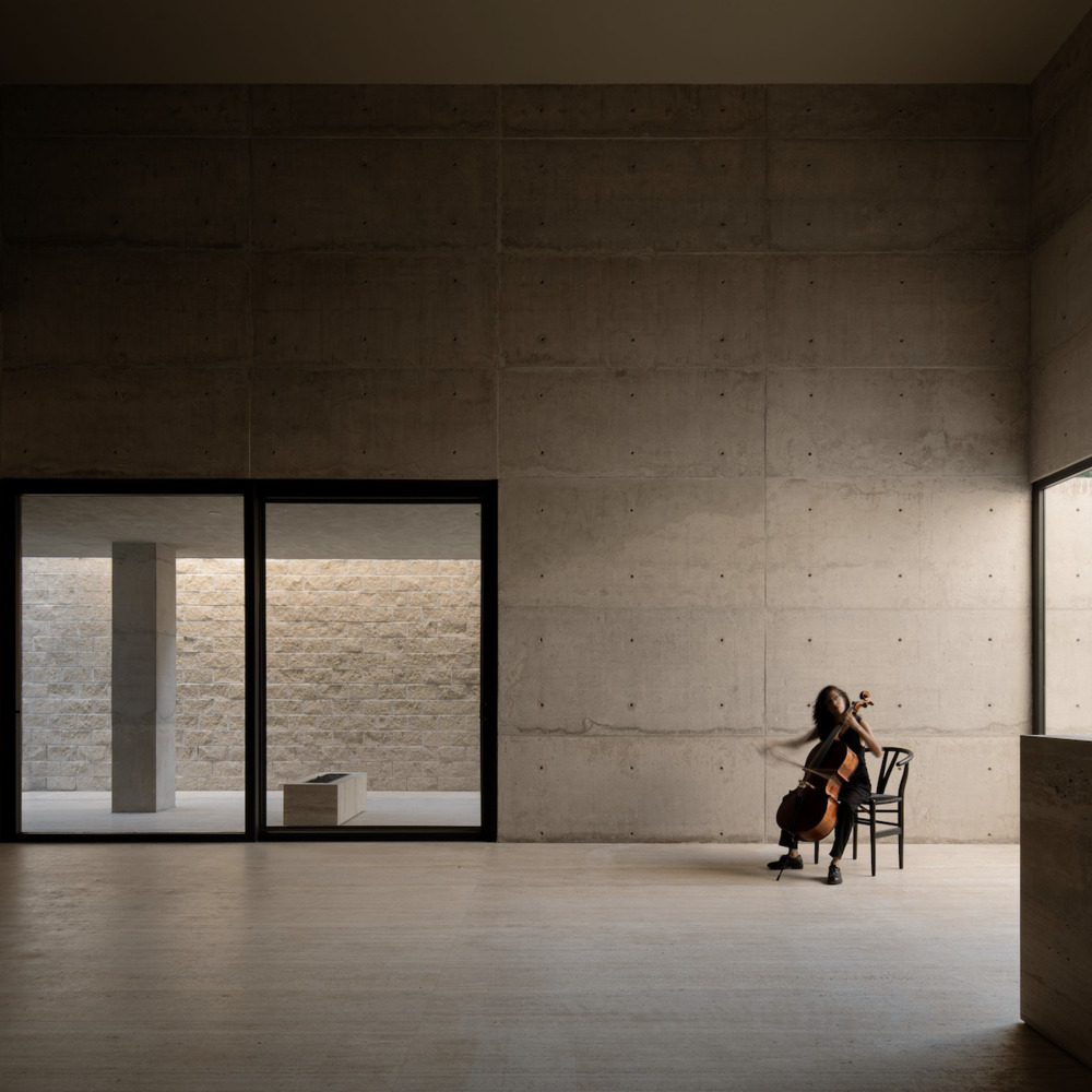 Board-formed concrete interior with cellist seated by floor-to-ceiling glazing overlooking a courtyard