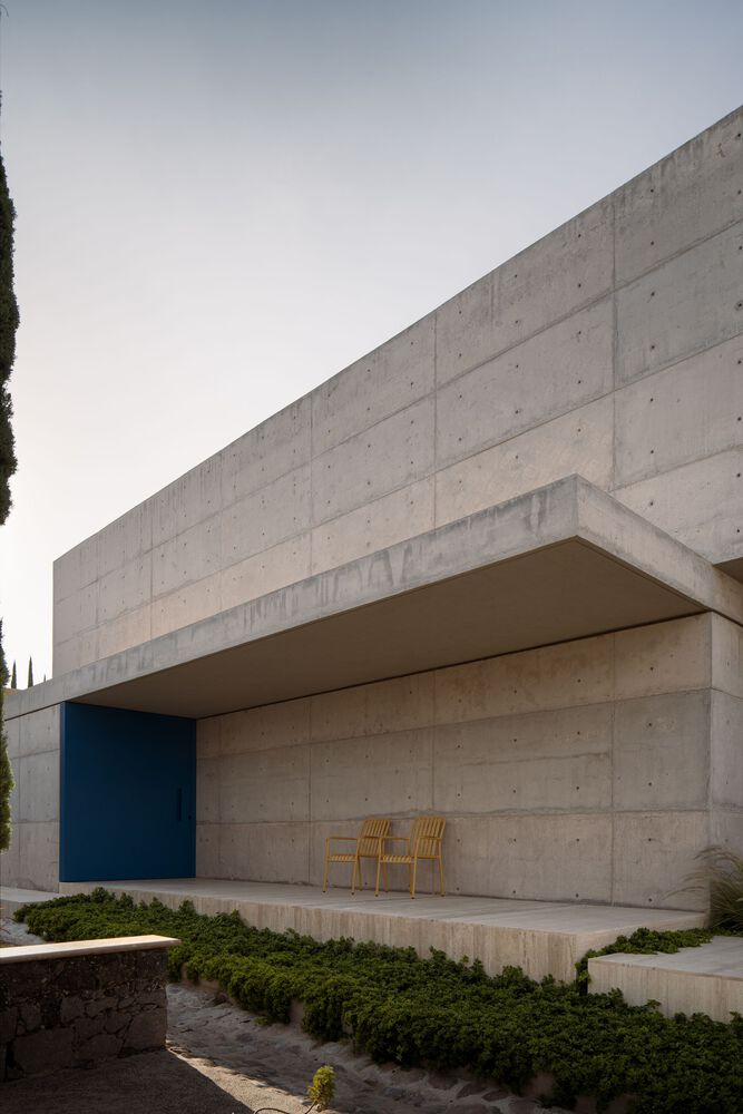 Cantilevered concrete overhang sheltering a recessed porch with two timber chairs and low planting
