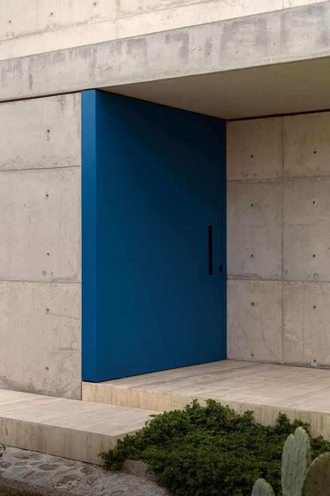 Blue pivot door set within board-formed concrete frame above planted bed with cacti and shrubs