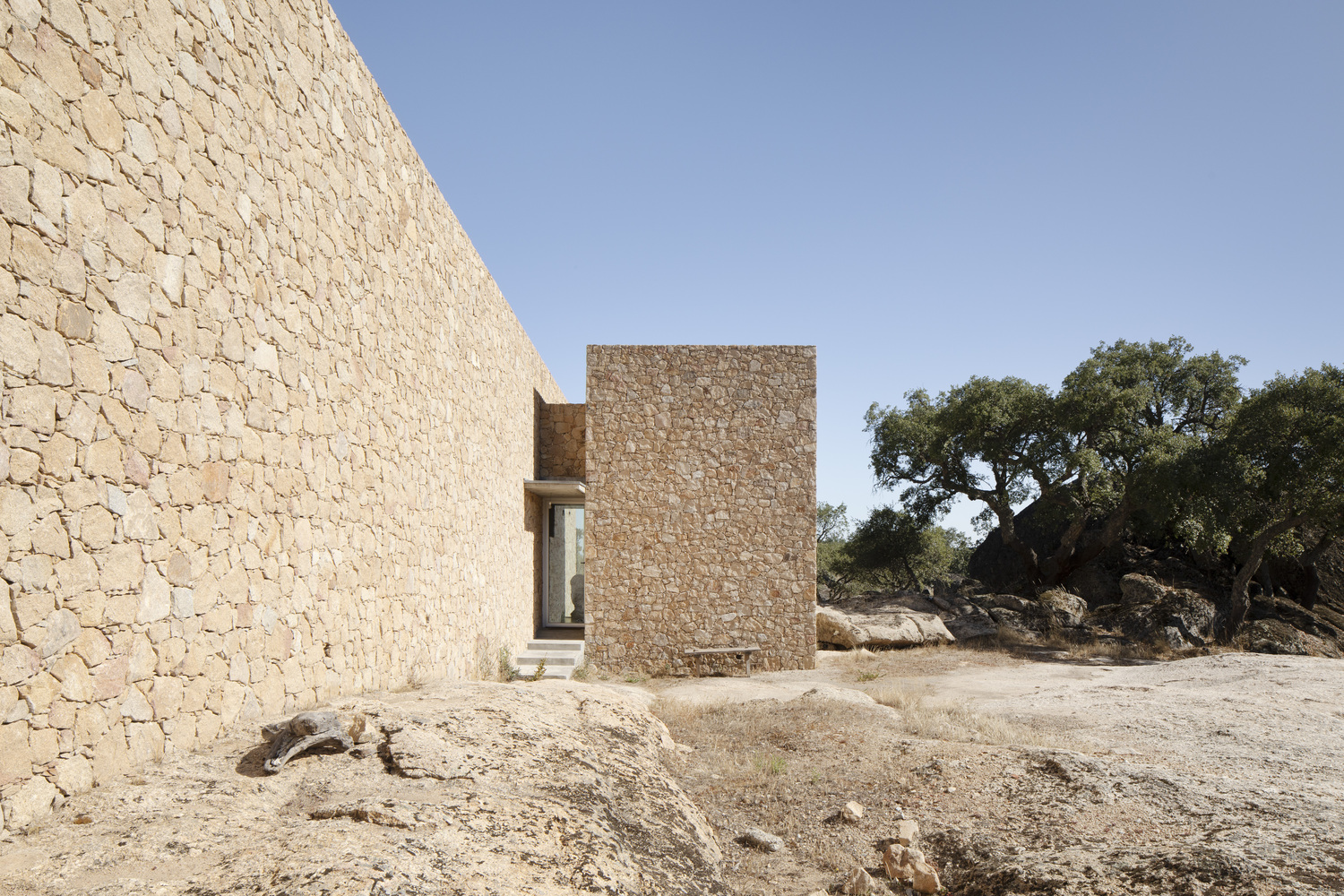 Stone facade volumes framing an entry passage with ancient olive trees and rocky terrain beyond