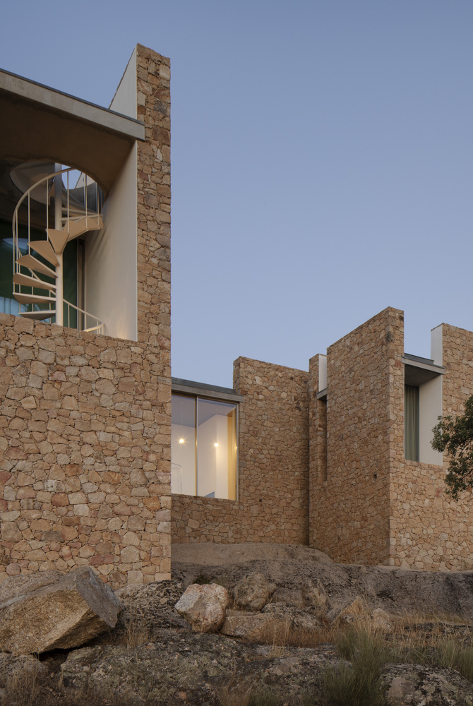 Stone walls and white volumes with a spiral staircase visible through an arched opening at dusk