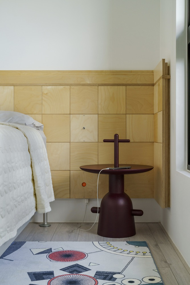 Bedroom corner with light wood paneled wall behind a dark sculptural side table and patterned rug