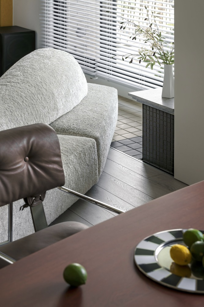 Living area with white horizontal blinds filtering daylight onto pale wood floors and tiled radiator cover
