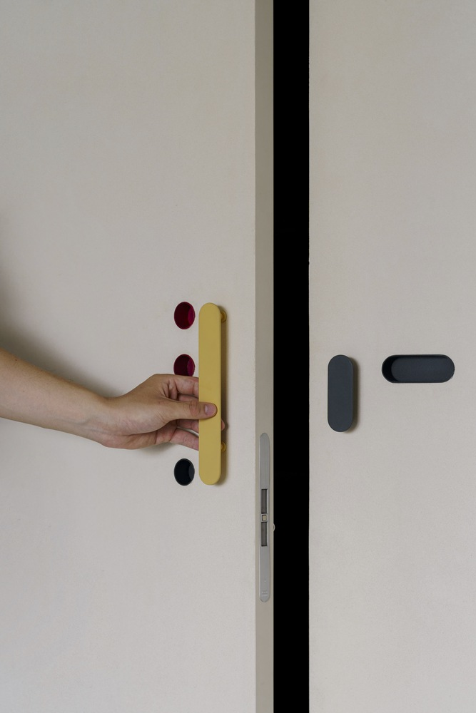 Hand operating yellow door handle with red and black accent buttons on white panel