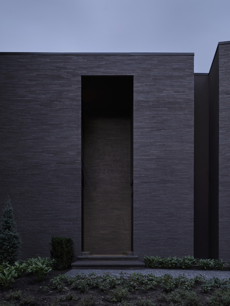 Dark brick facade with recessed timber entrance door flanked by low evergreen plantings at dusk