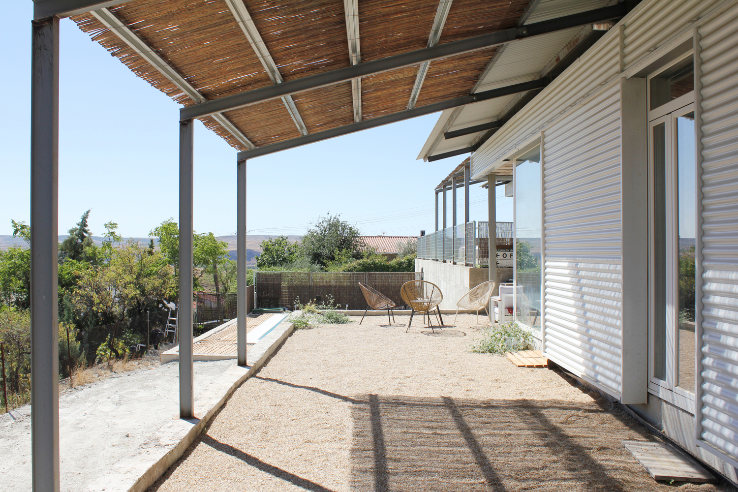 Covered terrace with timber slat pergola and steel columns casting shadows on gravel surface