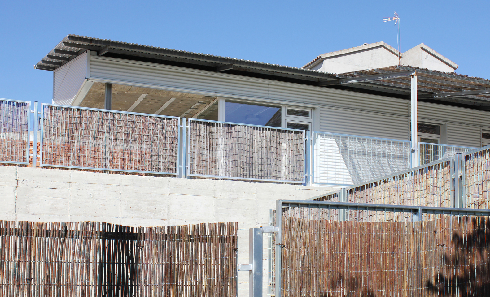 Street view of corrugated metal facade with bamboo screen balcony and concrete plinth under clear sky