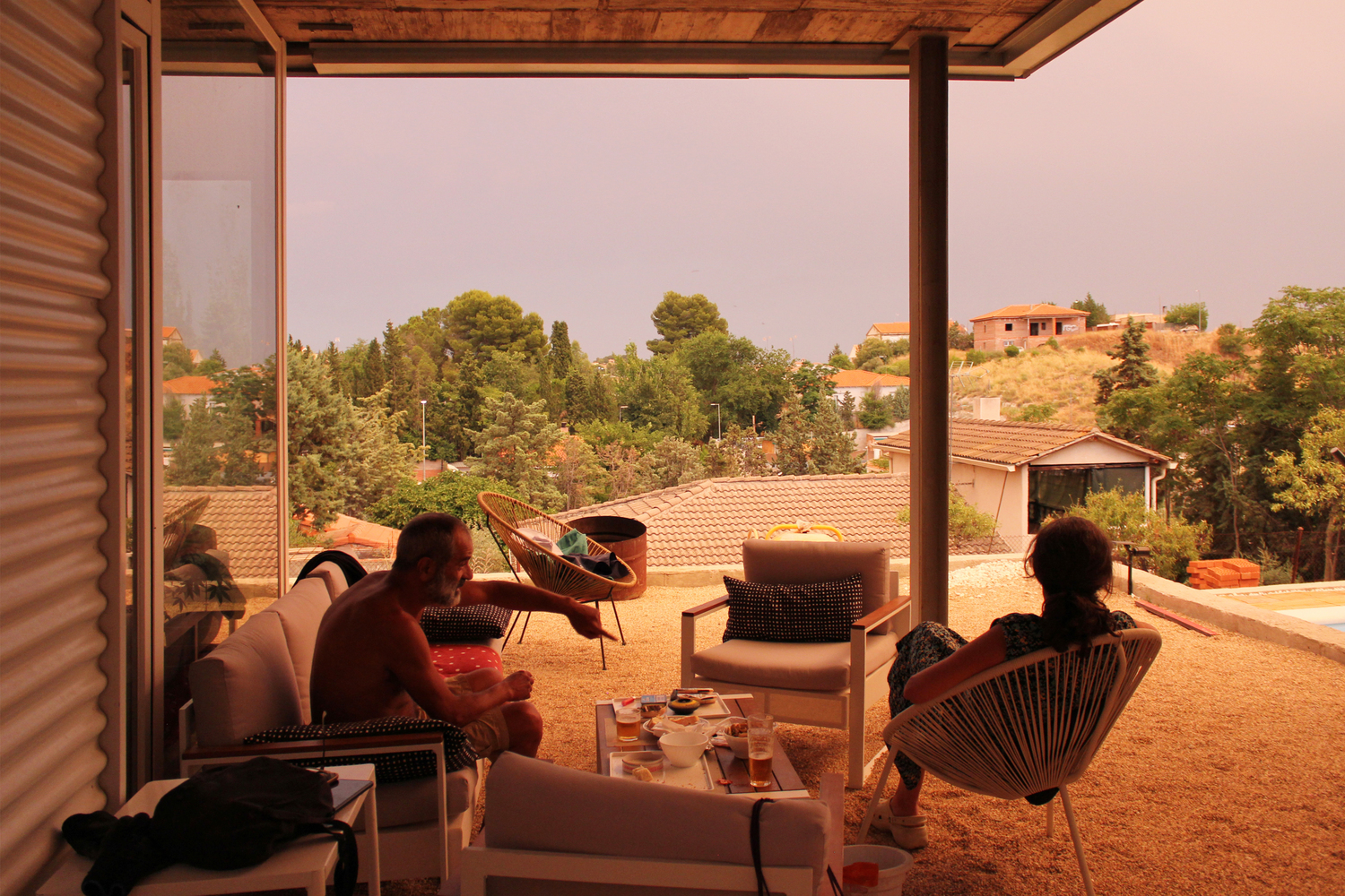 Covered terrace with timber pergola framing three seated figures overlooking tiled rooftops and hillside vegetation
