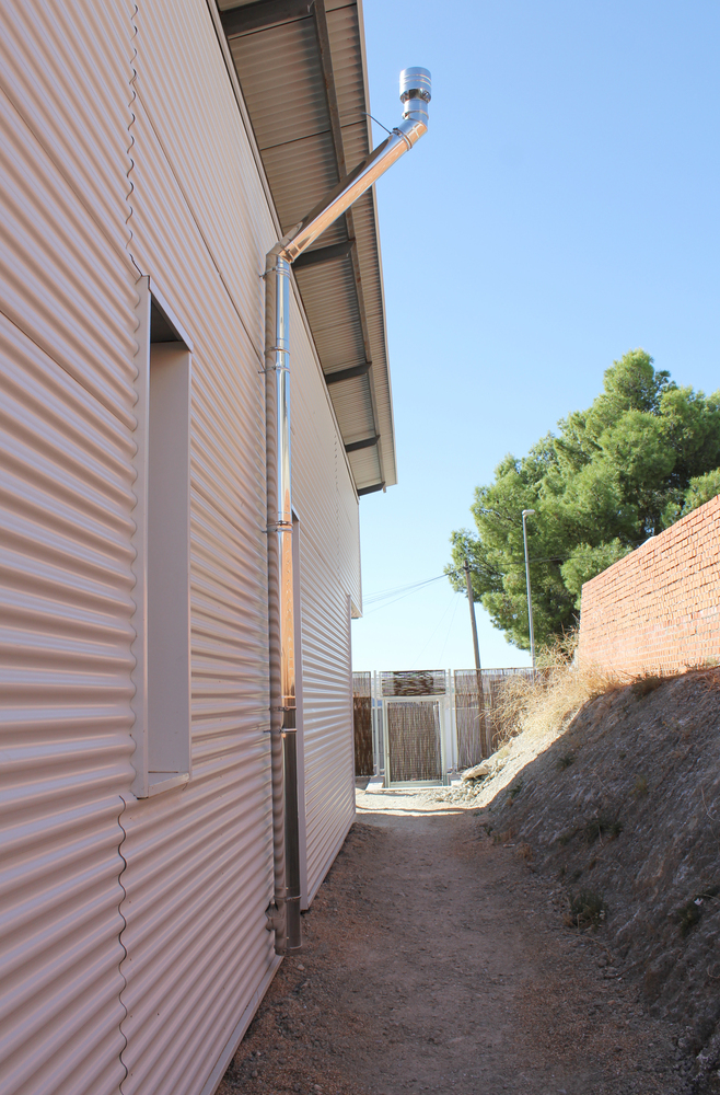 Narrow side passage between corrugated metal wall and sloped earth embankment with exposed drainage pipe