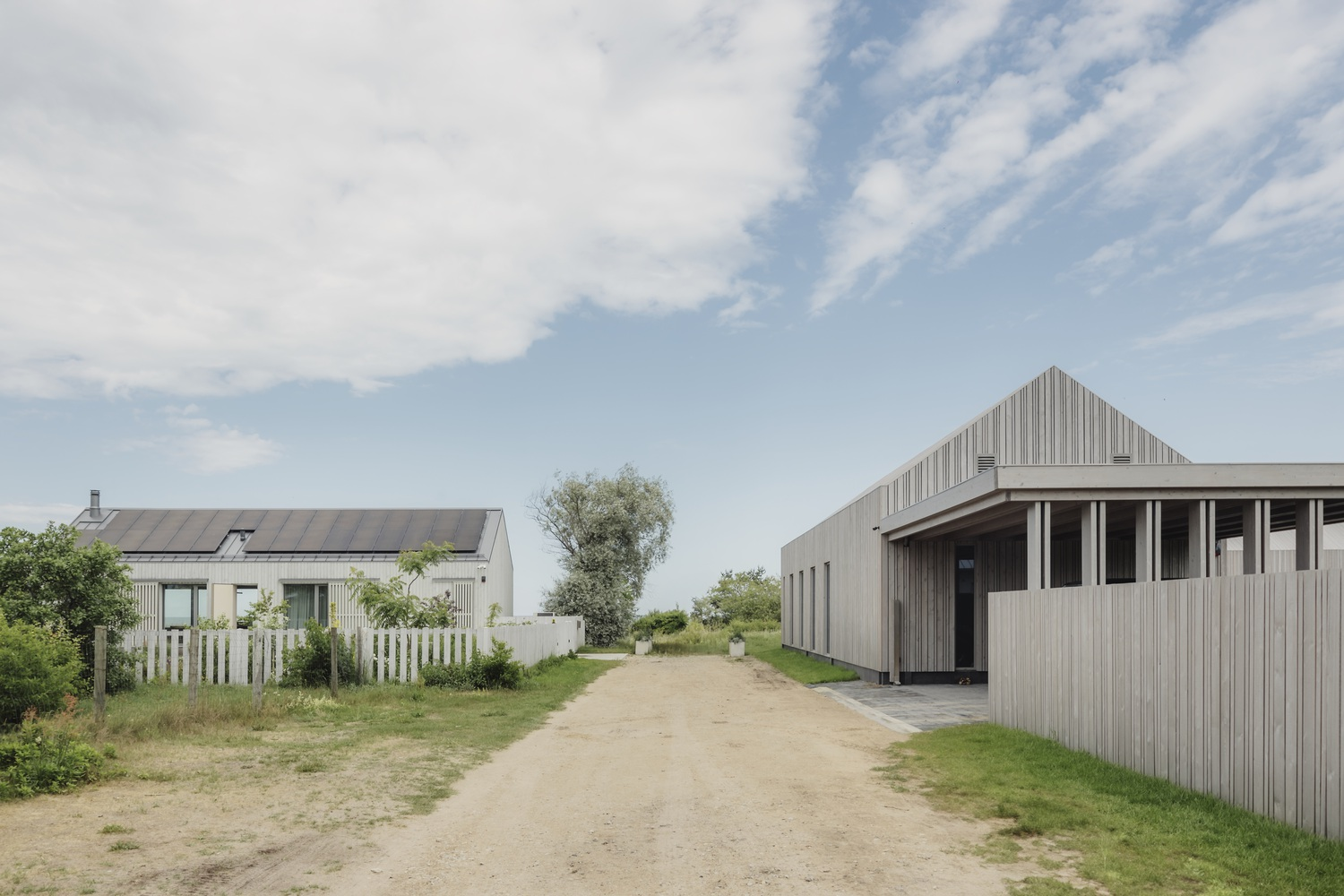 Gravel drive between two timber-clad gabled volumes under a partly cloudy sky with trees